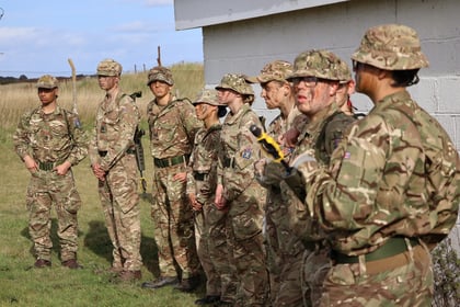 A group of soldiers in camouflage uniforms and hats stand outside near a white building, some with painted faces, listening attentively. The setting appears to be a grassy area with a fence and blue sky in the background.