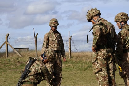Four soldiers in camouflage uniforms stand outdoors on grass near a wire fence, engaged in discussion or task. The sky is partly cloudy and they wear helmets and kit.