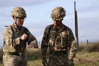 Two soldiers in camouflage uniforms and helmets are outdoors by a fence, focusing on handling a piece of black equipment or wire together. They stand on grass with a cloudy sky in the background.
