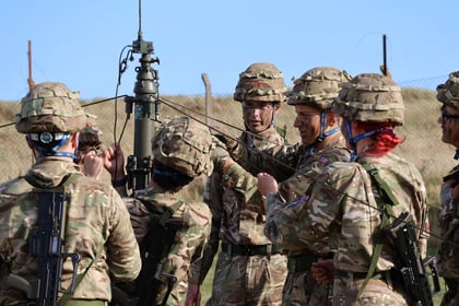 A group of soldiers in camouflage uniforms and helmets stand outdoors, gathered around a piece of military equipment, appearing to discuss or adjust it, with a clear sky in the background.