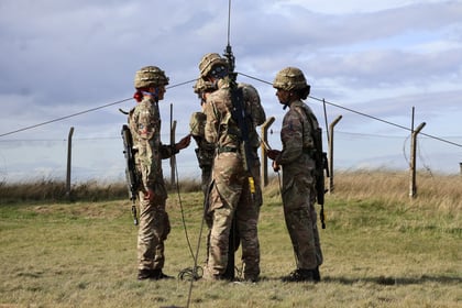 Four soldiers in camouflage uniforms and helmets stand in a circle outdoors, holding communication equipment, with a fence and cloudy sky in the background.