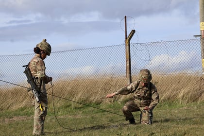 Two soldiers in camouflage uniforms work with wires on grassy ground near a barbed wire fence, under a cloudy sky.