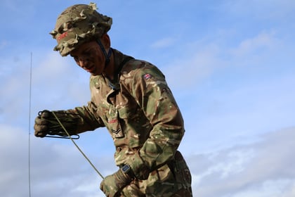 A soldier in camouflage uniform and helmet, holding a rope against a blue sky with clouds. The soldier's face is painted and he wears gloves and a watch.