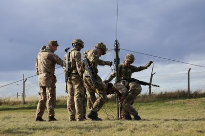 Four soldiers in camouflage uniforms set up a tall radio aerial outdoors on grassy terrain near a wire fence, working together under a cloudy sky.