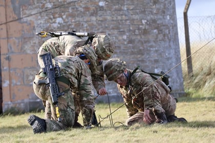 Three soldiers in camouflage uniforms kneel on grass, working with communications equipment outdoors near a stone building and a wire fence.
