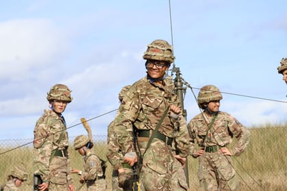 Several soldiers in camouflage uniforms and helmets stand outdoors, surrounded by wires and poles. The main person in the centre is smiling. A blue sky and a grassy field are visible in the background.