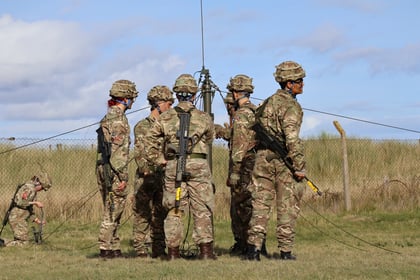 Seven soldiers in camouflage uniforms and helmets stand in a grassy field, gathered around communication equipment, with another soldier working in the background near a fence. Blue sky and clouds are visible.