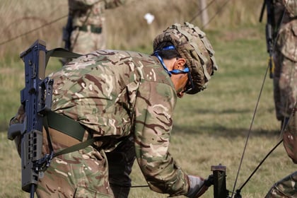 A soldier in camouflage uniform and helmet works with wires outdoors, appearing to set up equipment on grassy ground. Other soldiers are blurred in the background.