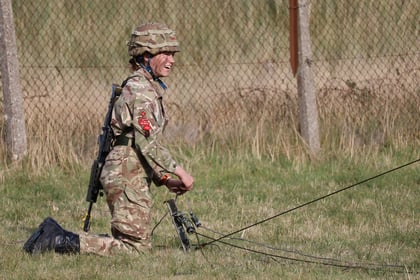 A person in military camouflage gear kneels on grass, smiling whilst handling communications equipment, with a rifle on their back and a fence in the background.