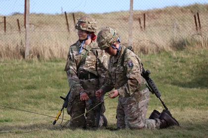 Two soldiers in camouflage uniforms and helmets kneel on grass, working with wires or equipment outdoors near a chain-link fence and tall grass.