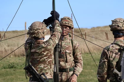 Three soldiers in camouflage uniforms set up radio equipment outdoors on a grassy field, with one adjusting wires whilst others observe. A fence and open sky are visible in the background.