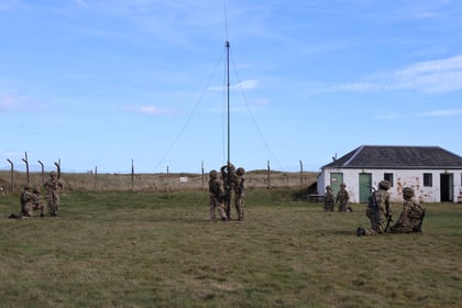 Several soldiers in camouflage uniforms work in groups on a grassy field, some near a tall aerial, with a small white building and a wire fence in the background under a blue sky.