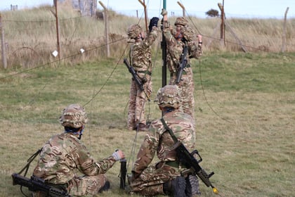 Four soldiers in camouflage uniforms set up communication equipment outdoors near a fence, working with cables and a tall pole on grassy terrain. Two are kneeling, and two are standing, all focused on the task.