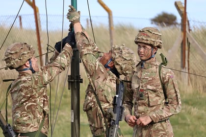 Three soldiers in camouflage uniforms set up equipment outdoors near a fence. One holds a pole while another adjusts it, and the third stands nearby, all with face paint and rifles. Grass and a wire fence are visible in the background.