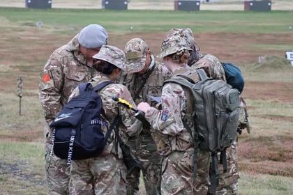 Five soldiers in camouflage uniforms and rucksacks gather closely together outdoors, looking down at a handheld device, possibly a map or satnav, in a grassy open field.