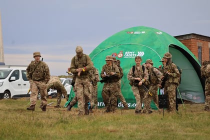 A group of army cadets in camouflage uniforms stand and walk near a green tent labelled Army Cadets on a grassy field with a van and brick building in the background.