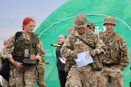 Three soldiers in camouflage uniforms walk outdoors; one carries a map, another carries a rifle. They are in front of a green tent, with a smiling person in the background. The sky is overcast.