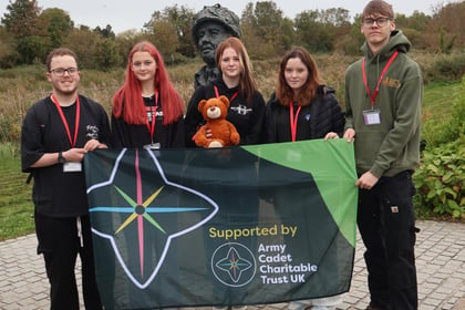 Four youths and one person in a green hoodie hold an Army Cadet Charitable Trust UK flag in front of a bronze statue head during the Normandy Battlefield Tour 2026.