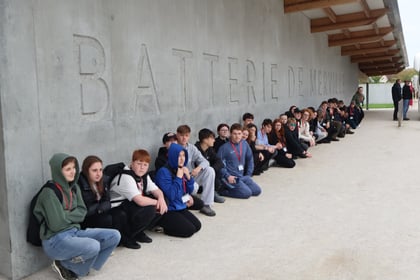 A large group of students on a Normandy Battlefield Tour 2026 sit in a row against a concrete wall inscribed with the words BATTERIE DE MERVILLE.