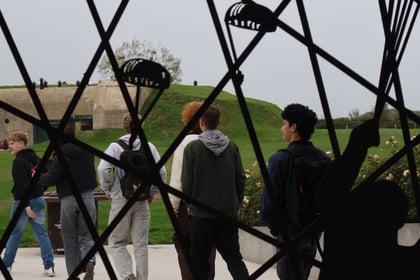 A group of students on a Normandy Battlefield Tour 2026 walks towards a grassy bunker, viewed through a metal gate decorated with silhouettes of aeroplanes and paratroopers.