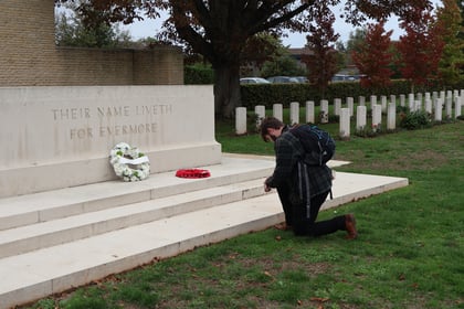 A young person with a rucksack kneels before a stone memorial inscribed with THEIR NAME LIVETH FOR EVERMORE, surrounded by the rows of headstones seen on a Normandy Battlefield Tour 2026, with a wreath resting on the steps.