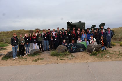 A group of students and chaperones on a Normandy Battlefield Tour 2026 stand with lanyards before a parked tank, two people in front holding a banner that says Supported by Army Benevolent Fund.