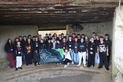 A large group of teenagers stands together inside a concrete bunker during a Normandy Battlefield Tour 2026, holding a green banner that reads Supported by Army Cadet Charitable Trust UK.