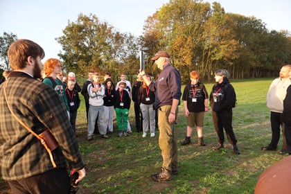 A man in a baseball cap stands in a field holding up a clipboard and speaking to a group of young people wearing red lanyards during a Normandy Battlefield Tour 2026.