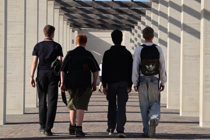 Four people walk away from the camera through a stone memorial corridor lined with pillars engraved with names, evocative of a Normandy Battlefield Tour 2026.
