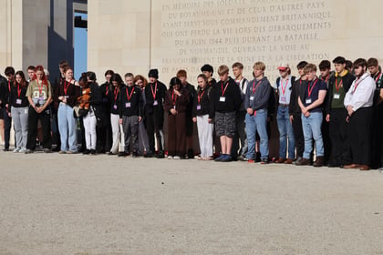 A large group of students stands with heads bowed in front of a stone memorial wall inscribed with French text during a Normandy Battlefield Tour 2026.