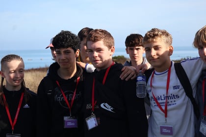 A group of teenagers wearing Normandy Battlefield Tour 2026 lanyards posing for a photo outdoors near the sea.