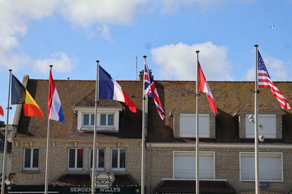 Six flags, including those of Belgium, France, the United Kingdom, and the United States, fly on poles in front of a brick building with a Sergeant Willys sign, a stop on a Normandy Battlefield Tour 2026.