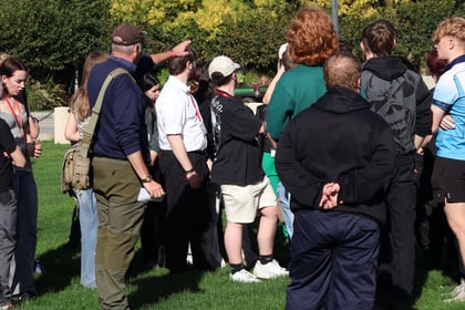 A group of people standing on a grassy field during a Normandy Battlefield Tour 2026, with one man pointing towards the distance.