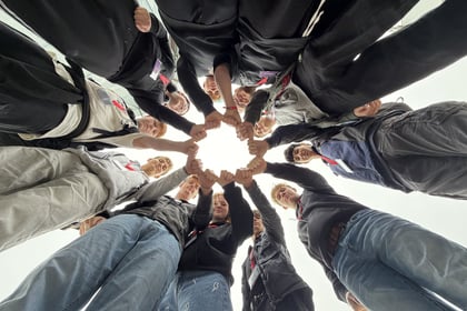 A group of teenagers on a Normandy Battlefield Tour 2026 stand in a circle looking down at the camera, joining their fists together in the centre.