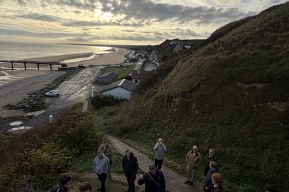 A group on a Normandy Battlefield Tour 2026 stands on a grassy hillside path, listening to a guide whilst overlooking a coastal town, beach, and pier under a cloudy sky.