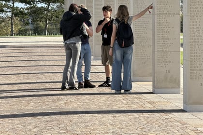 Four people stand in an outdoor memorial colonnade, with one person pointing to a name inscribed on a stone pillar during a Normandy Battlefield Tour 2026.