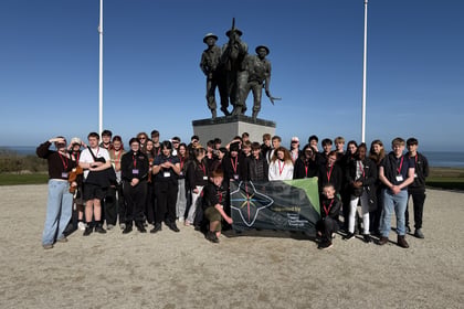 A large group of teenagers in casual clothing pose before a bronze war memorial statue during a Normandy Battlefield Tour 2026, with one student kneeling in front holding a flag that reads Supported by Army Cadet Charitable Trust UK.