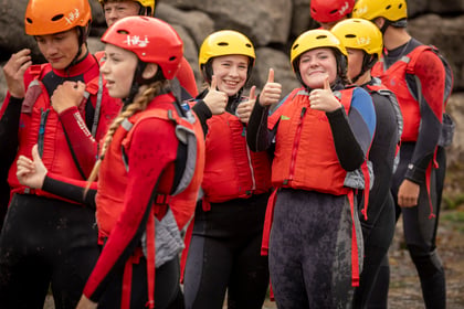 A group of people in wetsuits, red life jackets, and helmets stand outdoors, smiling. Two people in the centre are giving thumbs up, whilst others appear to be chatting or preparing for an activity.