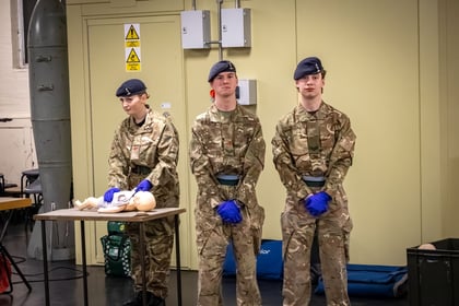 2 ACF cadets stand in MTP uniform, hands clasped infront of them wearing medical gloves. To the side is another cadet dressed the same but with a first aid baby dummy in front of her on a table.