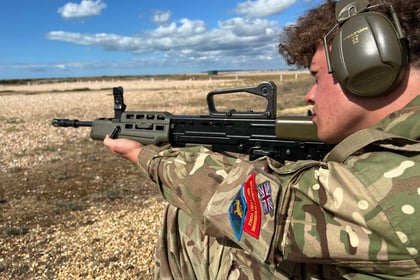 A person in camouflage uniform and ear protection aims a rifle outdoors on a rocky field under a blue sky with clouds. A patch with the Union Flag and a red emblem is visible on the sleeve, suggesting participation in the Berkshire ACF Summer Camp 2026.