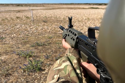 Person in camouflage uniform holding and aiming a rifle at an outdoor shooting range during the Berkshire ACF Summer Camp 2026, with a clear sky and open field in the background.