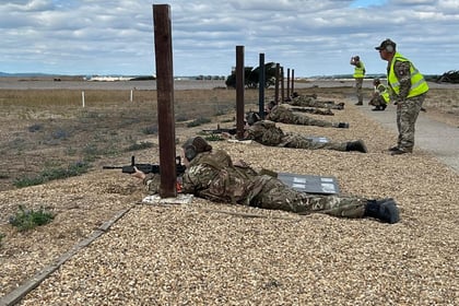Several people in military uniforms lie on the ground aiming rifles at targets, while two individuals in yellow vests stand nearby on a gravel range under a partly cloudy sky during Berkshire ACF Summer Camp 2026.