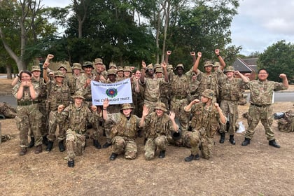 A large group of people in camouflage uniforms smile and cheer outdoors, holding up a banner reading The Connaught Trust. Trees and dry grass are in the background, capturing the lively spirit of the Berkshire ACF Summer Camp 2026.