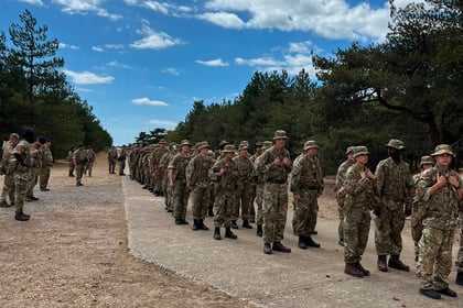 A group of soldiers in camouflage uniforms stand in formation on a wide path outdoors, surrounded by trees under a partly cloudy blue sky during the Berkshire ACF Summer Camp 2026.