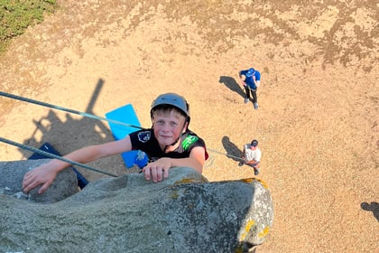 A person wearing a helmet climbs a rock wall, reaching the top and looking up during the Berkshire ACF Summer Camp 2026. Three people stand on the ground below, watching. The scene is outdoors on a sunny day.