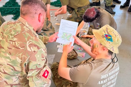 A group of people in military uniforms sit and kneel on the floor, examining maps and papers during the Berkshire ACF Summer Camp 2026. One person holds a map, whilst others look on or work together, engaged in a collaborative group activity.