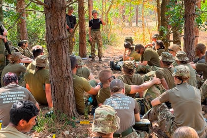 A group of cadets in camouflage uniforms sit in a forest, listening to an instructor standing by the trees. Sunlight filters through the leaves, and recruitment details are visible on some T-shirts, marking the energetic spirit of Berkshire ACF Summer Camp 2026.