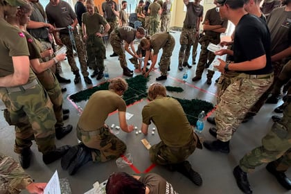 A group of people in camouflage uniforms gather indoors around a large map on the floor at the Berkshire ACF Summer Camp 2026, some kneeling or standing, discussing and pointing at the map, with kit and papers scattered nearby.