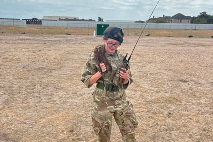 A person in camouflage military uniform and boots stands on dry grass, holding a radio with a long aerial, outdoors under a cloudy sky with buildings and a fence in the background—capturing the spirit of Berkshire ACF Summer Camp 2026.