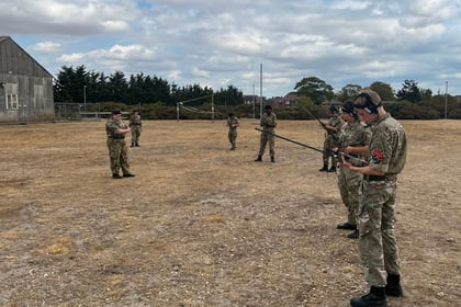 Several people in military uniforms stand spaced out on a dry, grassy field under a partly cloudy sky, with one person appearing to address the group. A building and trees are visible in the background, capturing a moment from Berkshire ACF Summer Camp 2026.